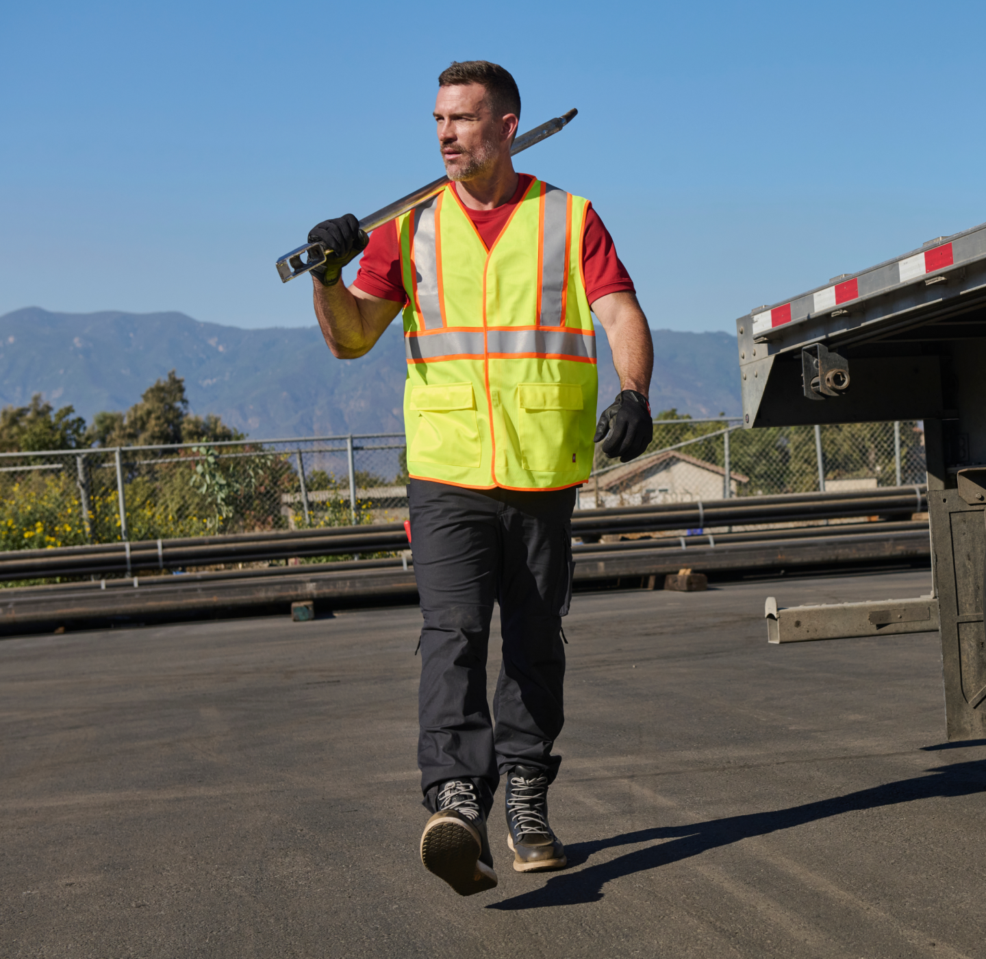 A man in a yellow safety vest carries a large wrench on his shoulder while walking near a flatbed truck in an industrial outdoor setting, with mountains in the background.