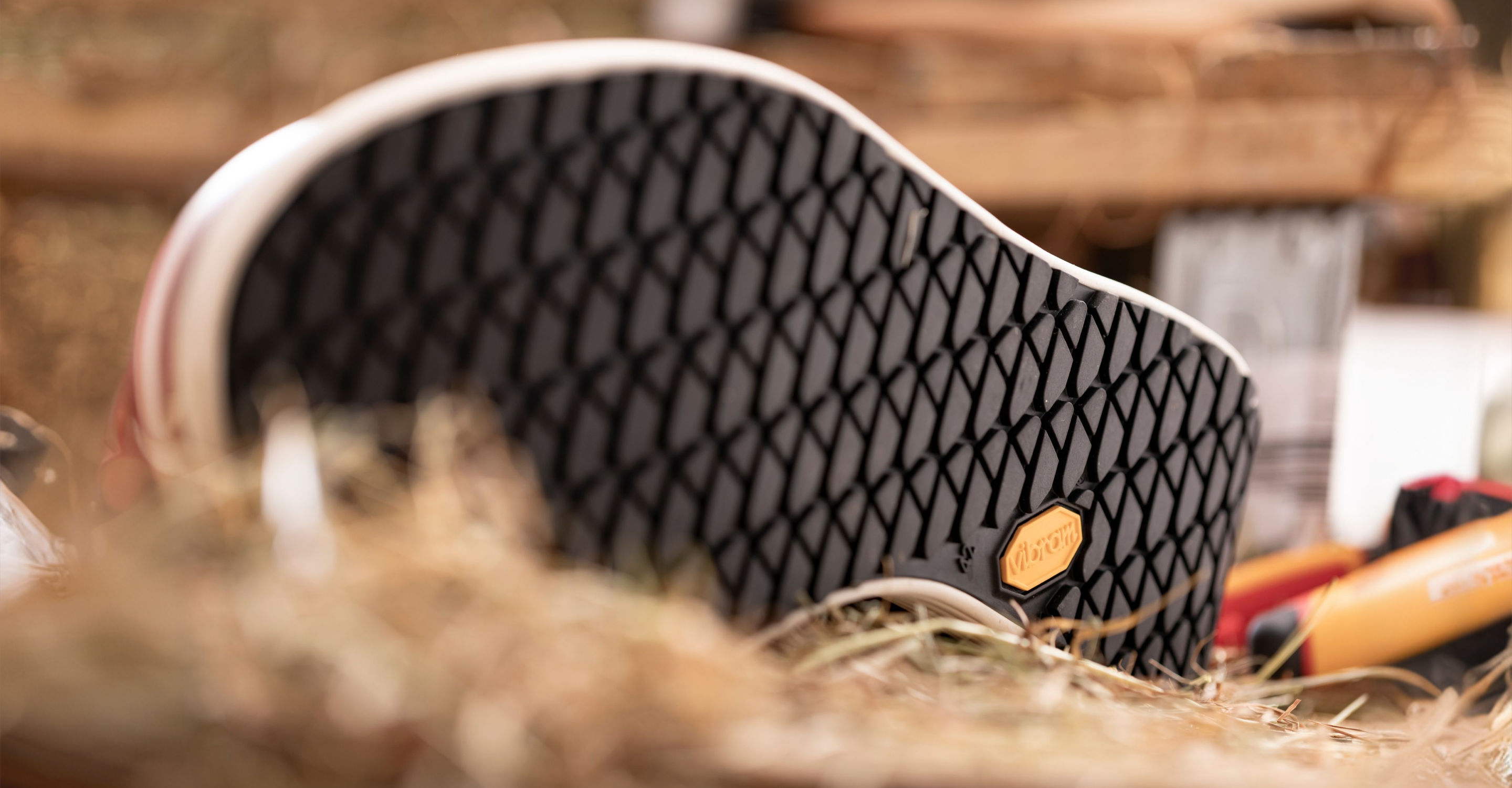 Close-up of a black shoe sole with a diamond pattern and a yellow hexagonal logo, lying on hay with blurred wooden objects in the background.