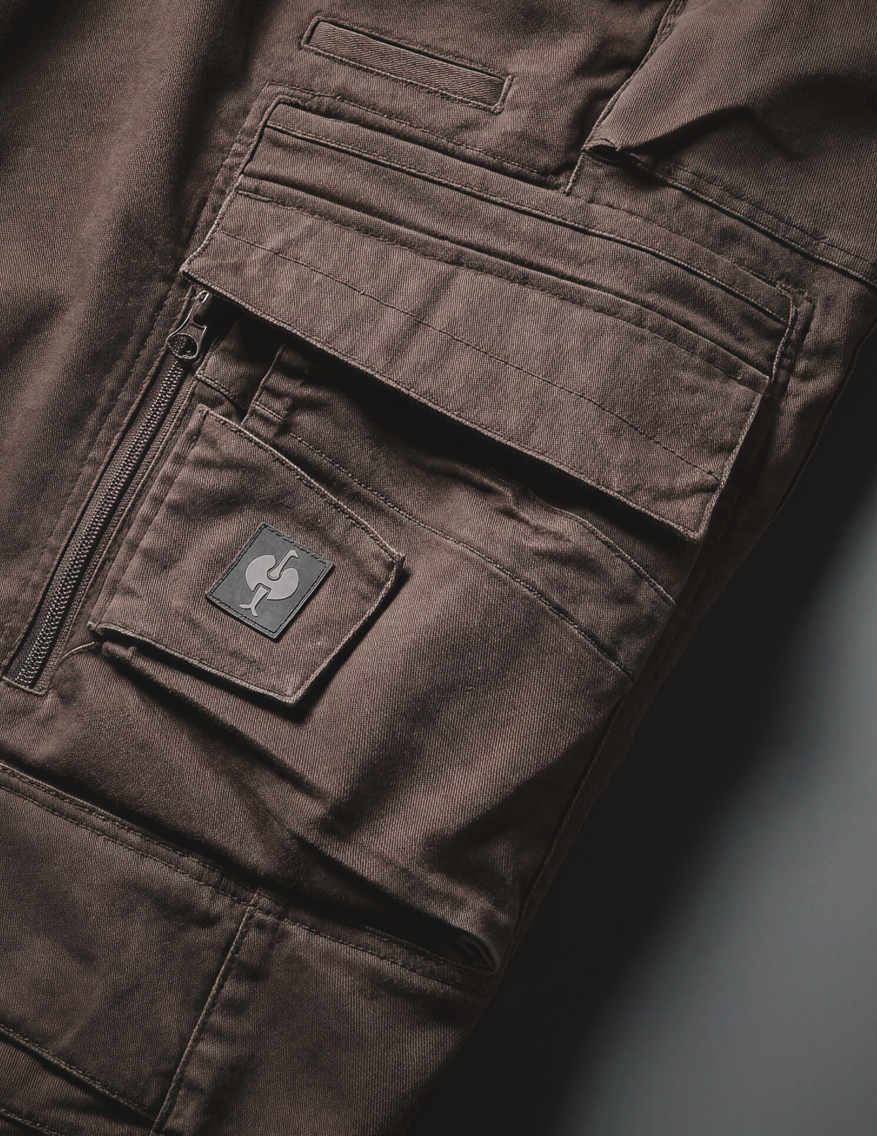 Close-up of the pocket of a brown cargo work pants with a visible brand logo on a gray background