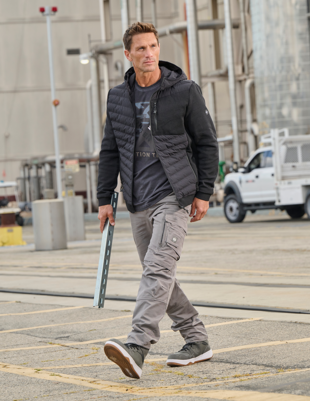 Man walking outdoors in a parking lot wearing a black jacket and gray pants.