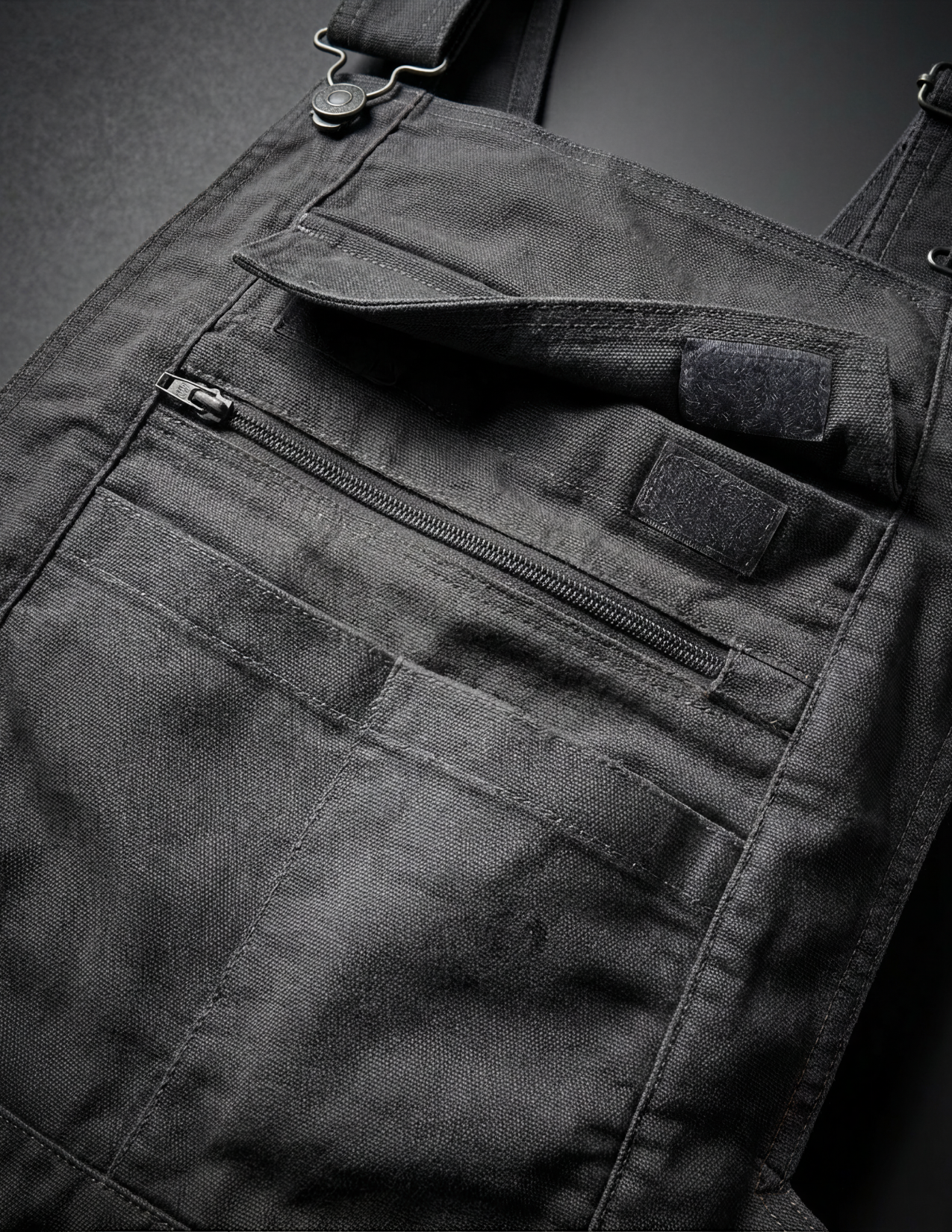 Close-up of dark gray bib overalls with a zipper pocket on a black background