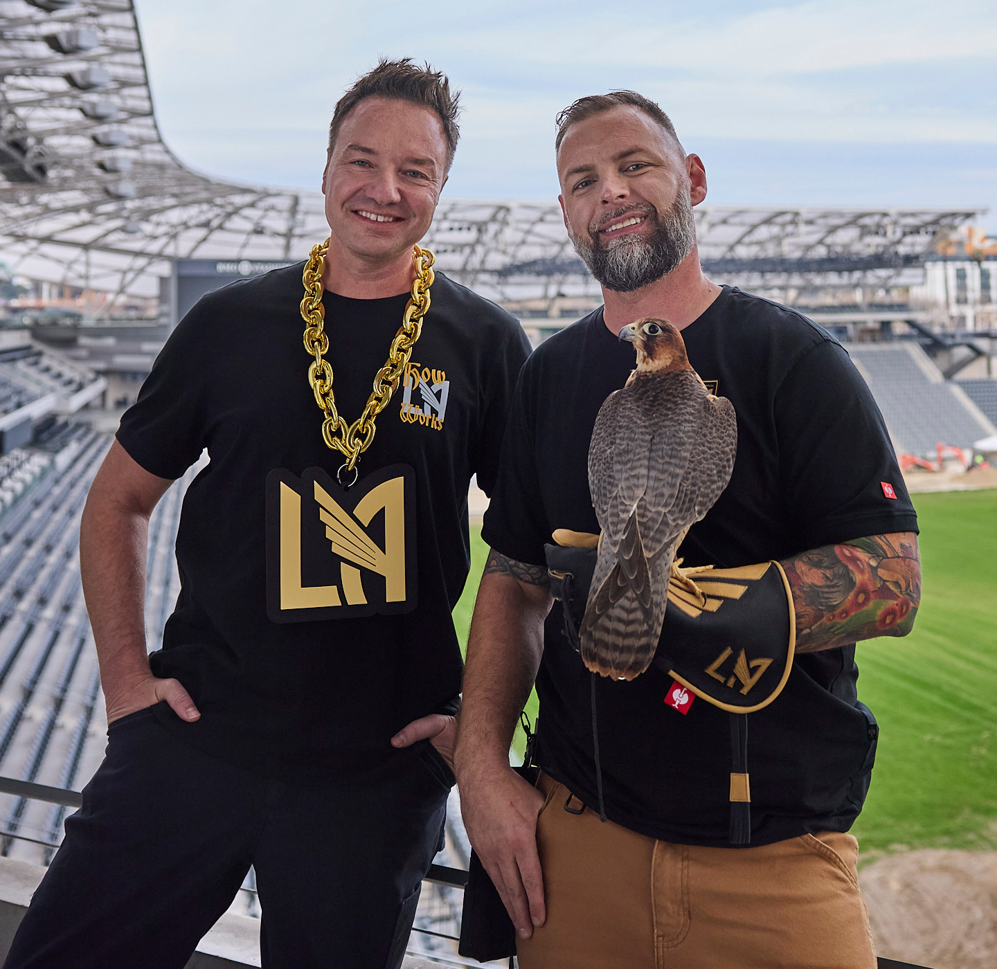 Two men stand in a stadium wearing LAFC shirts. One has a large gold LAFC chain, while the other holds a falcon wearing a glove. The stadium seats and field are visible in the background.