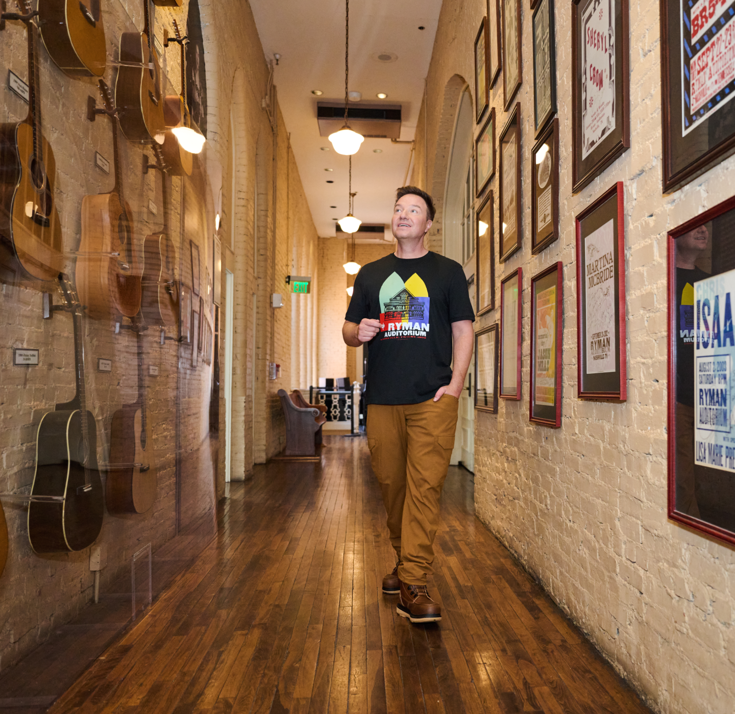 A man walks down a hallway with wooden floors, framed posters on one wall, and guitars displayed on the other, looking up thoughtfully.