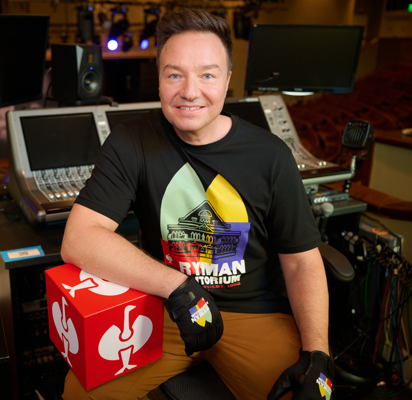 Smiling person in a Ryman Auditorium shirt and gloves sits at a soundboard, with one arm on a red box showing a white logo. Stage lights and empty seats are visible in the background.