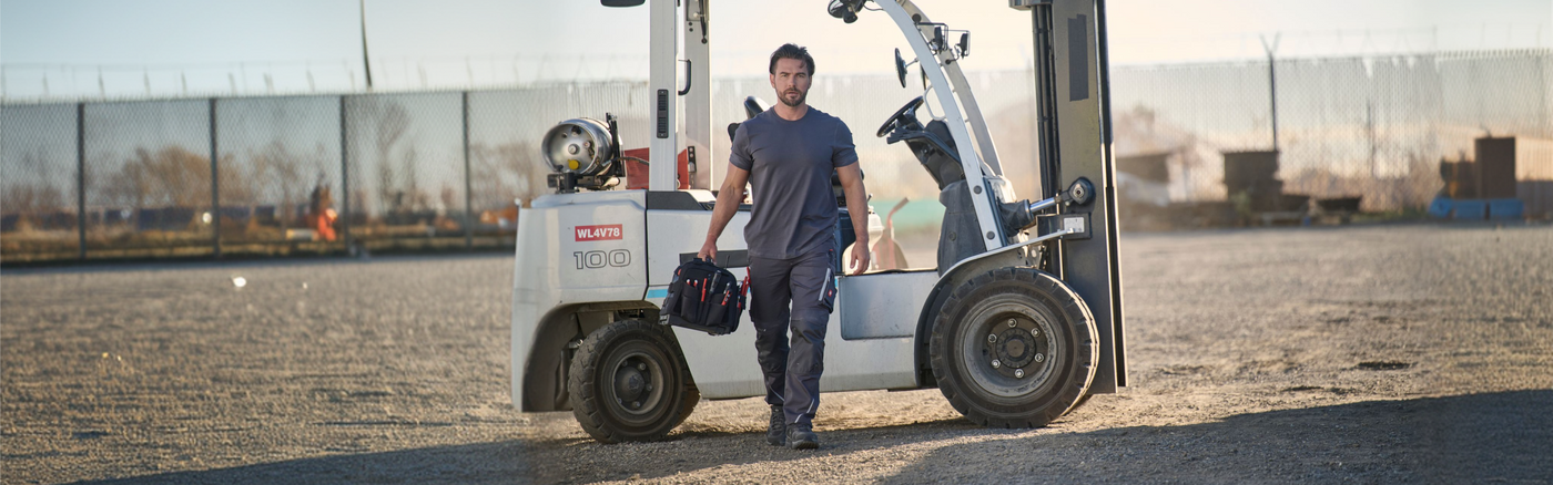 A man holding a bag with tools in his hand, walking infront of a forklift