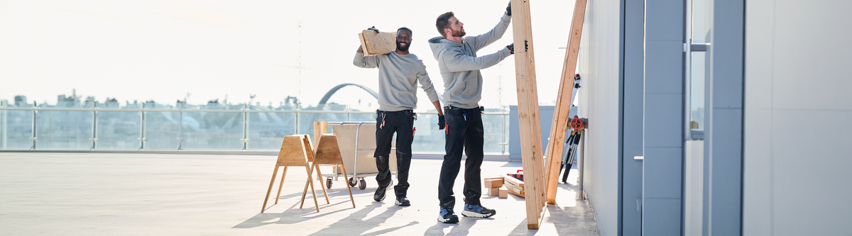 Two men in work clothes handle lumber on a rooftop. One carries wooden planks, the other secures a board to a structure. Tools and sawhorses are nearby, with a cityscape blurred in the background.