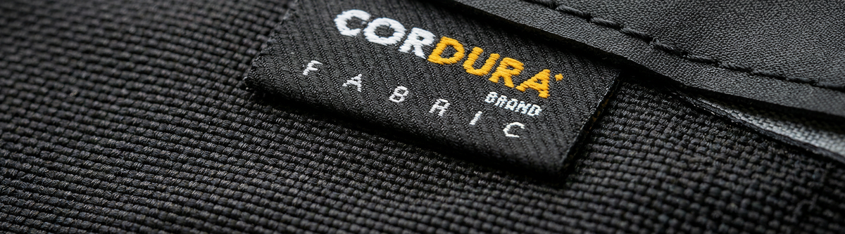 Close-up of a black CORDURA® brand fabric label stitched onto textured black material, with the words CORDURA BRAND FABRIC visible in white and yellow text.