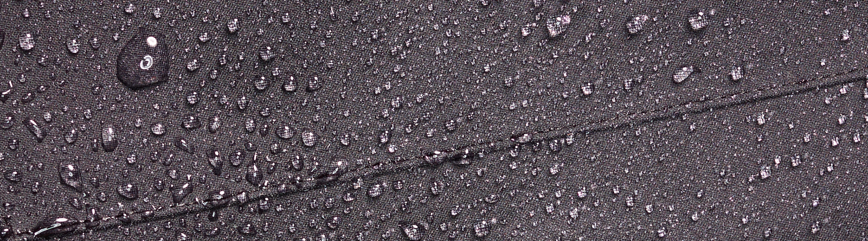 Water droplets scattered on a dark gray fabric surface, showing its water-repellent properties. The fabric features a visible diagonal seam running across the image.