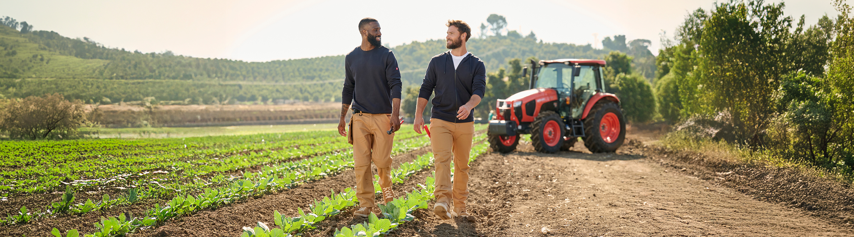 Two men walk and talk beside rows of crops on a sunny farm, with a red tractor parked on a dirt road in the background and green hills in the distance.