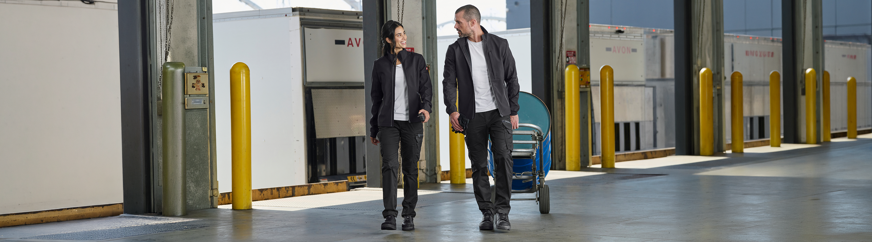 Two people in uniforms walk and talk in a warehouse loading dock area, with yellow bollards, rolling doors, and large containers in the background.