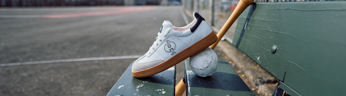 A white sneaker with a brown sole rests on a tennis ball, propped against a green bench on an empty outdoor sports court.