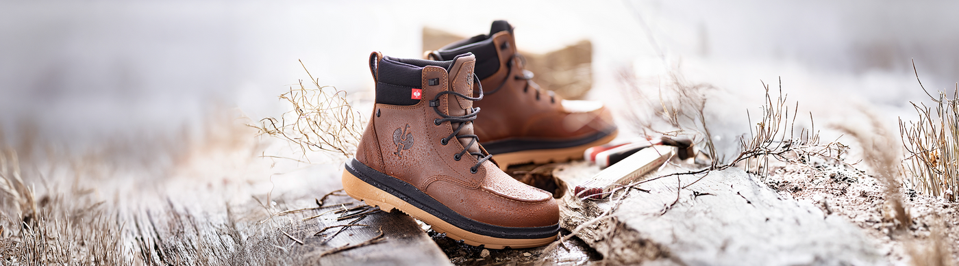 A pair of brown leather hiking boots with black soles sits on a rustic wooden surface outdoors, surrounded by dry grass and blurred natural elements in the background.