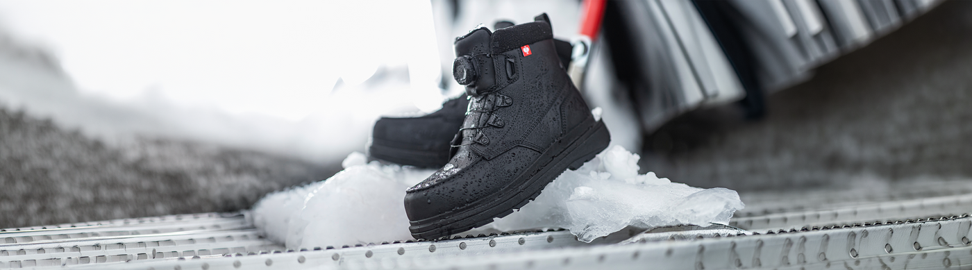 A pair of black winter boots with laces sits on ice-covered metal steps, with a blurred background suggesting a cold, outdoor environment.