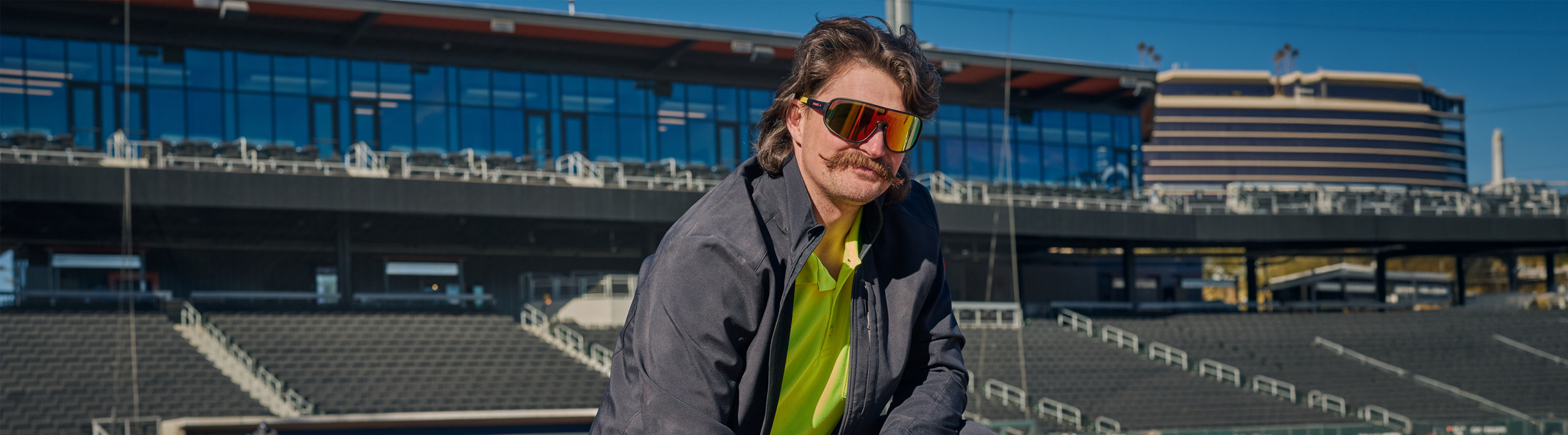 A man with a mustache and sunglasses wearing a gray jacket and neon green shirt poses in front of empty stadium seats and a modern building under a clear blue sky.