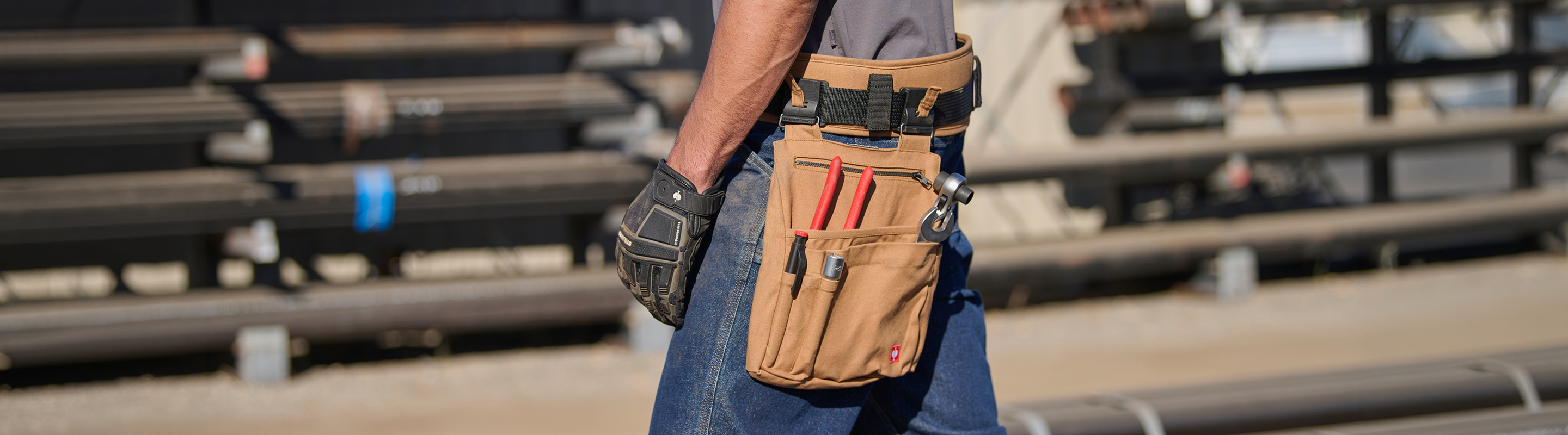 A person wearing a tool belt with various tools, including pliers and screwdrivers, stands outdoors near industrial pipes, wearing gloves and jeans.