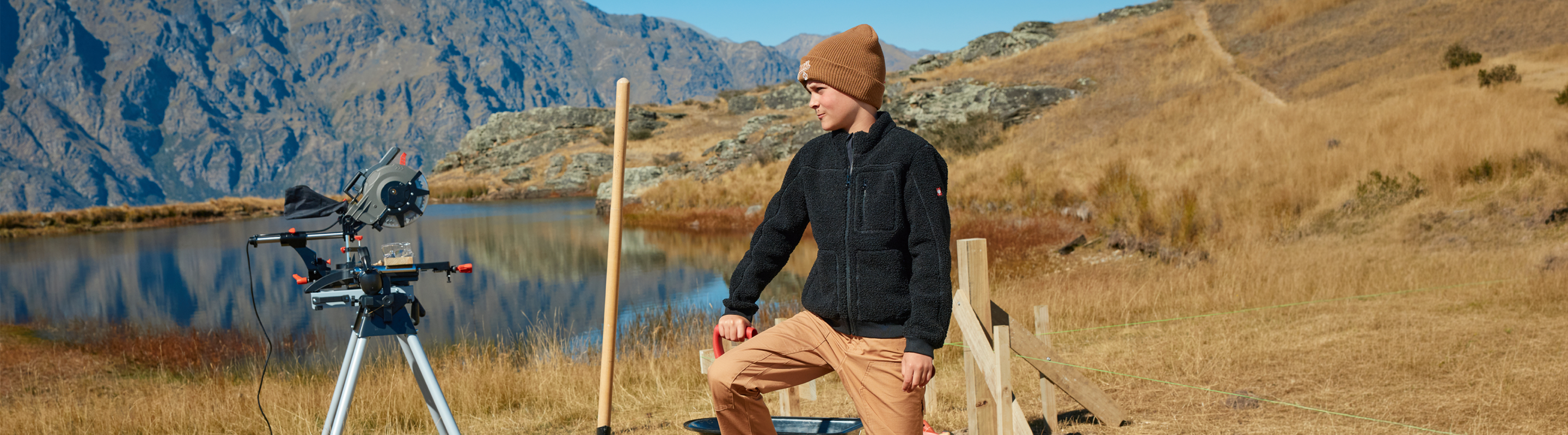 A person in a brown beanie and black jacket sits on a wooden fence near a lake, with mountains in the background and camera equipment set up on a tripod beside them in a grassy, outdoor area.