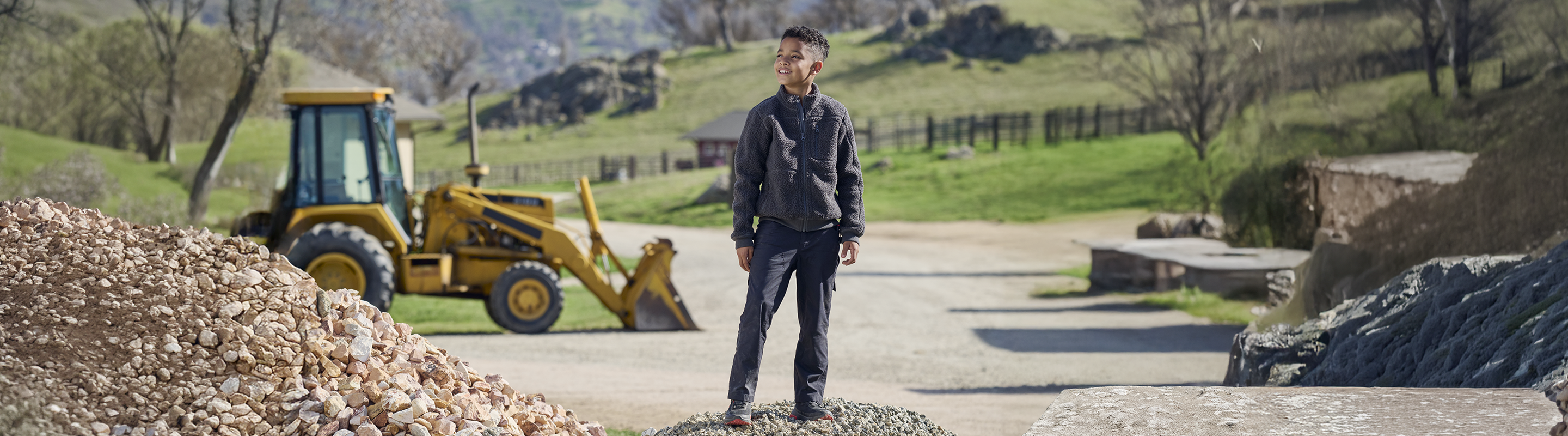 A boy stands on gravel at a construction site, with a yellow backhoe and green hills in the background on a sunny day.