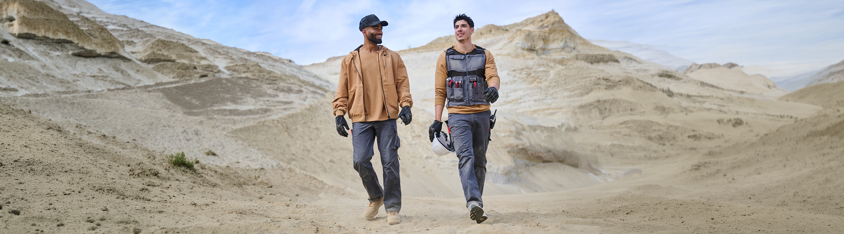 Two people in work clothes and gloves walk in a rocky desert landscape, with beige hills and blue sky in the background. One carries a vest and tools, and both appear to be talking and enjoying the scenery.