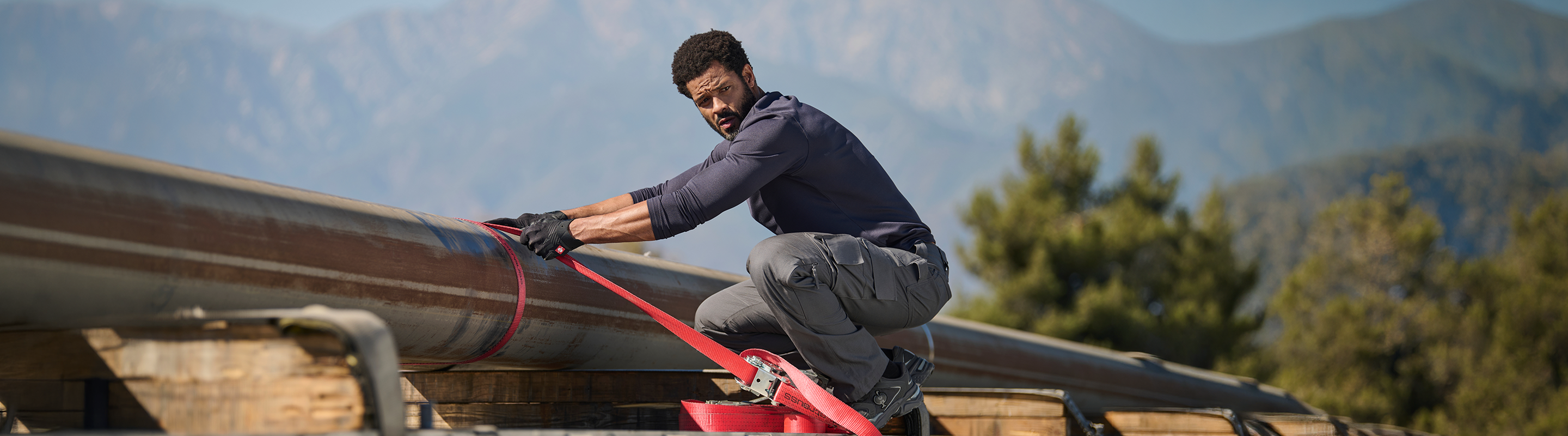 A person wearing gloves crouches on top of a large pipeline outdoors, pulling a red strap. Mountains and trees are visible in the background.
