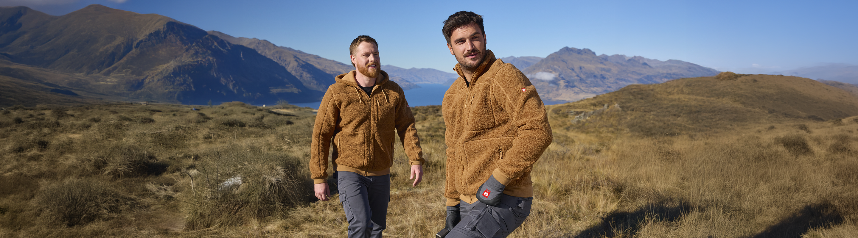 Two men standing outdoors in a mountainous grassy landscape, wearing matching brown fleece jackets. The background features rugged hills and mountains under a clear blue sky.