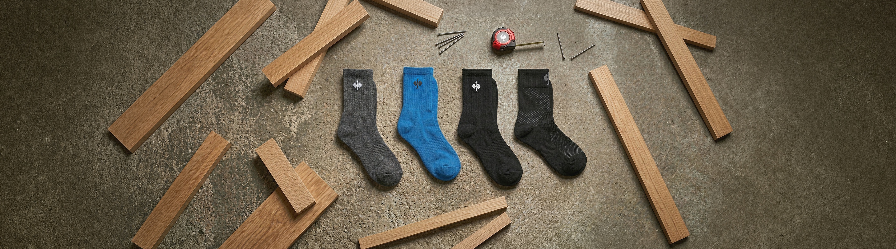 Five pairs of socks—gray, blue, and black—are lined up on a concrete floor, surrounded by scattered wooden planks, screws, and a tape measure.