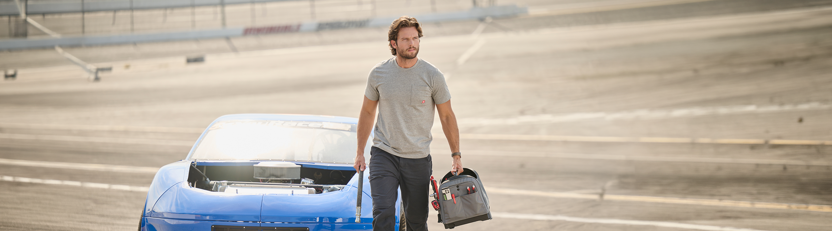 A man in a gray t-shirt and dark pants stands on an empty racetrack next to a blue car with its hood removed, holding a black tool bag.