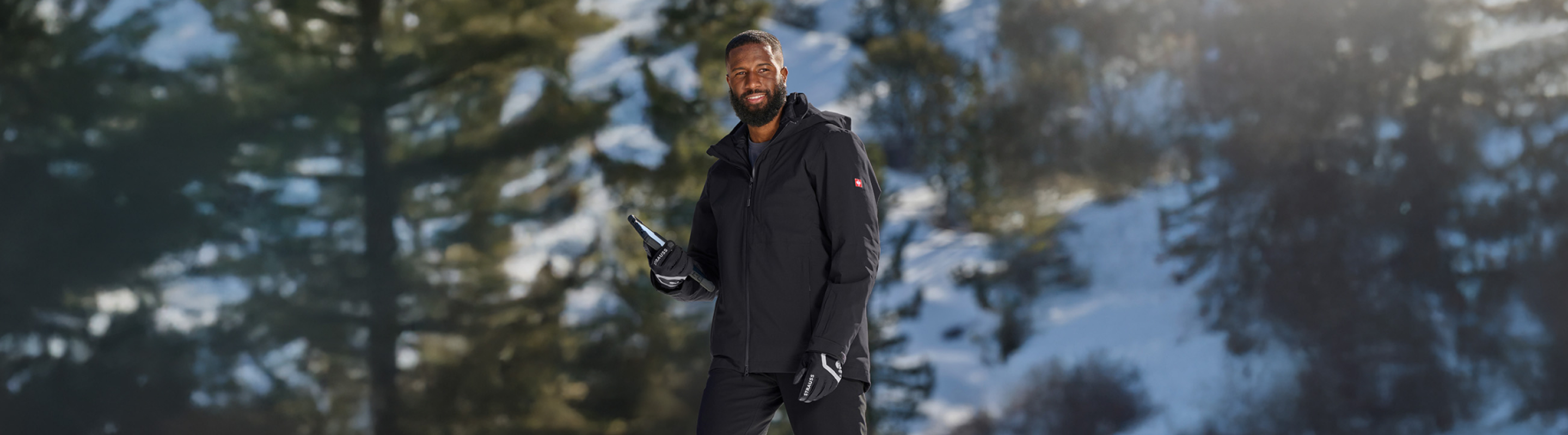 A man in a black jacket and gloves smiles while holding a phone, standing outdoors with snow and pine trees in the background.