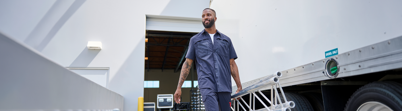 A man in a blue work uniform walks beside a large silver cargo container in a bright industrial setting.