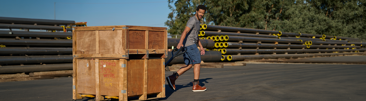 A man in a gray t-shirt and shorts walks beside a large wooden crate in an outdoor warehouse or yard, with stacks of black pipes in the background under a blue sky.