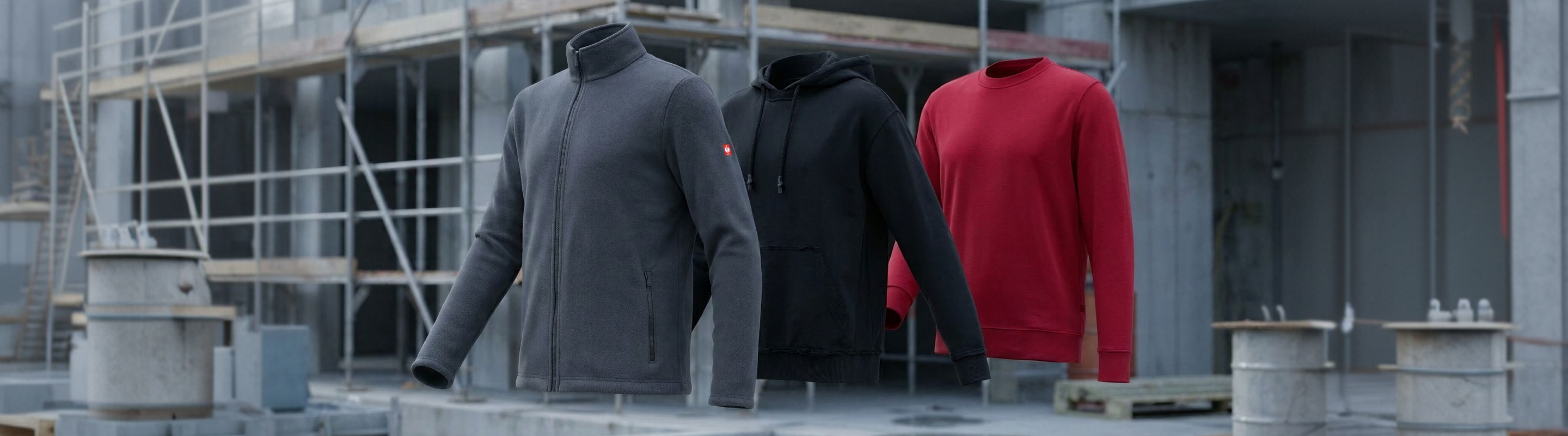 Three floating jackets—one gray zip-up, one black hoodie, and one red crewneck—are displayed in front of a building under construction with scaffolding and concrete structures.