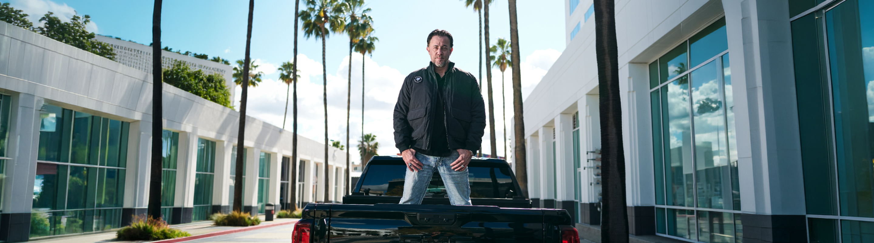 A man stands on the back of a black pickup truck parked between modern buildings and tall palm trees, with blue sky and clouds in the background.