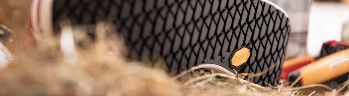 Close-up view of a black shoe sole with a textured pattern and a small yellow logo, set outdoors with dry grass and blurred gear in the background.