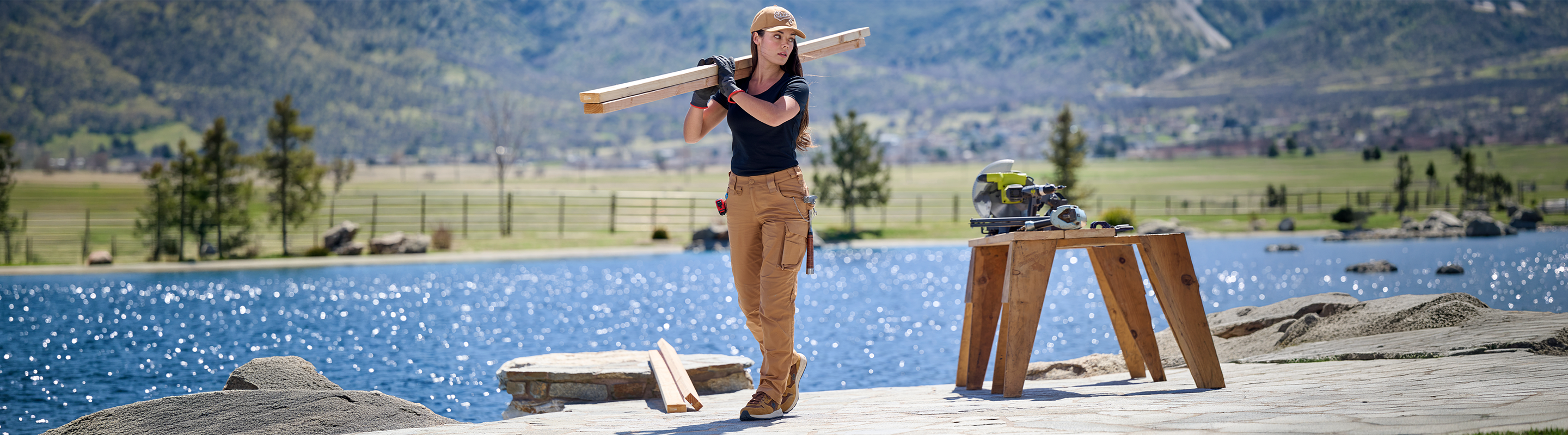 A woman stands on a wooden dock by a bright blue lake with mountains and trees in the background, holding a long wooden board over her shoulder. A small wooden table beside her holds boots and other gear.