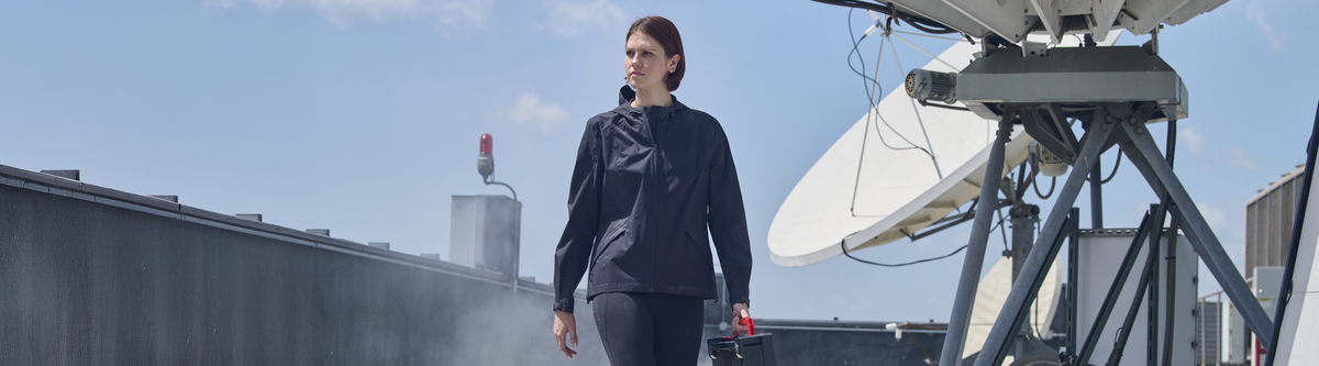 A woman in a dark navy long-sleeve athletic top and matching pants walks on a rooftop beside a large white satellite dish under a blue sky.