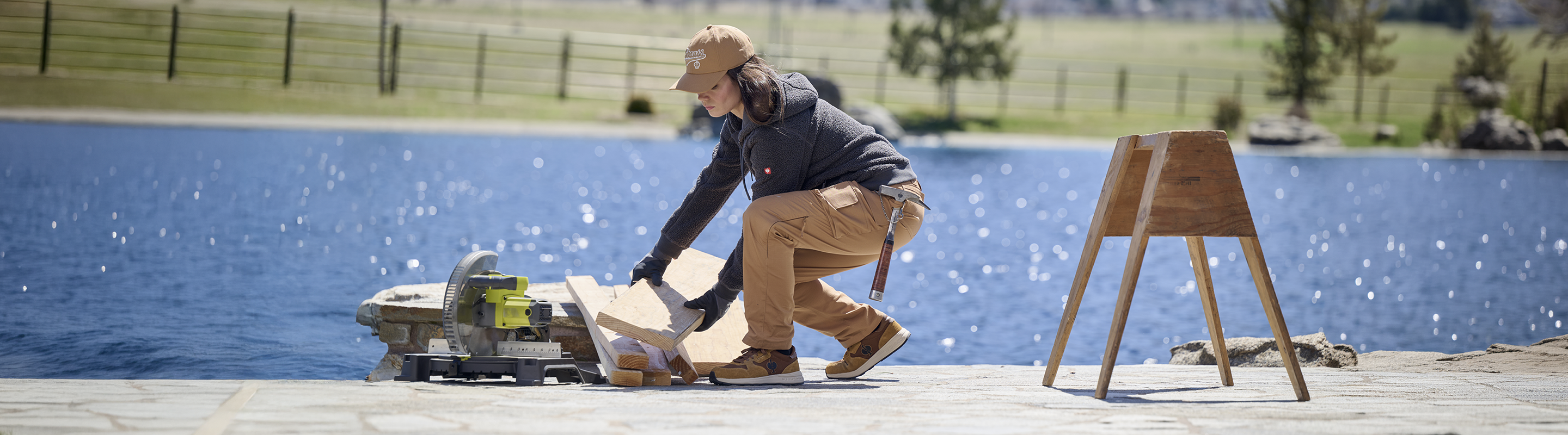 A person in work clothes kneels by a lake, using a table saw to cut wood. A wooden sawhorse stands nearby, and there is a scenic, fenced field in the background.