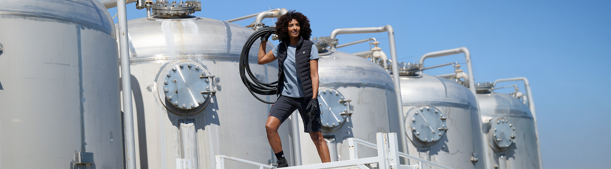 A man with curly hair stands on white metal walkways in front of large stainless steel tanks under a clear blue sky, holding a coiled black hose. No brand is visible.
