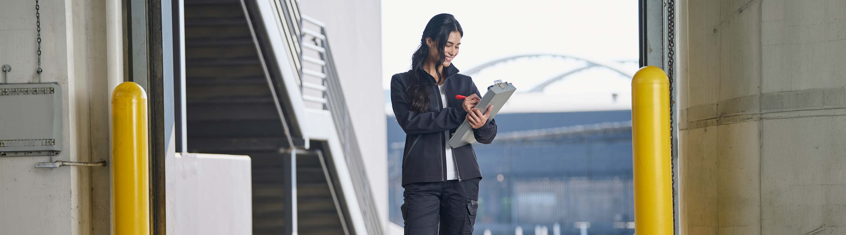 A woman stands between yellow posts outdoors, holding a clipboard and pen, and appears to be checking or recording information. There are stairs and industrial buildings in the background.