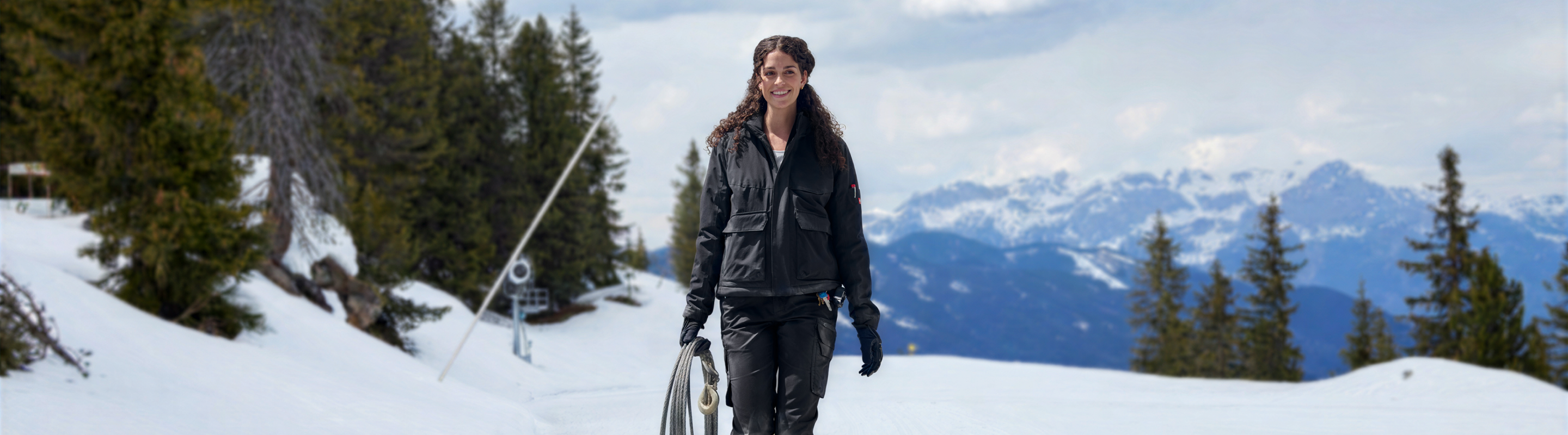 A woman in a black winter jacket and black pants walks through a snowy mountain landscape with pine trees and distant snow-capped peaks under a cloudy sky.
