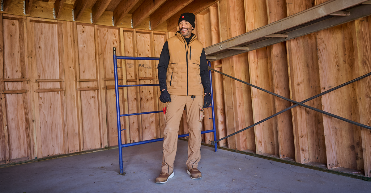 A man standing in a wooden room wearing a black beanie, sweatshirt, gloves and almondbrown vest, work pants and safety shoes. 