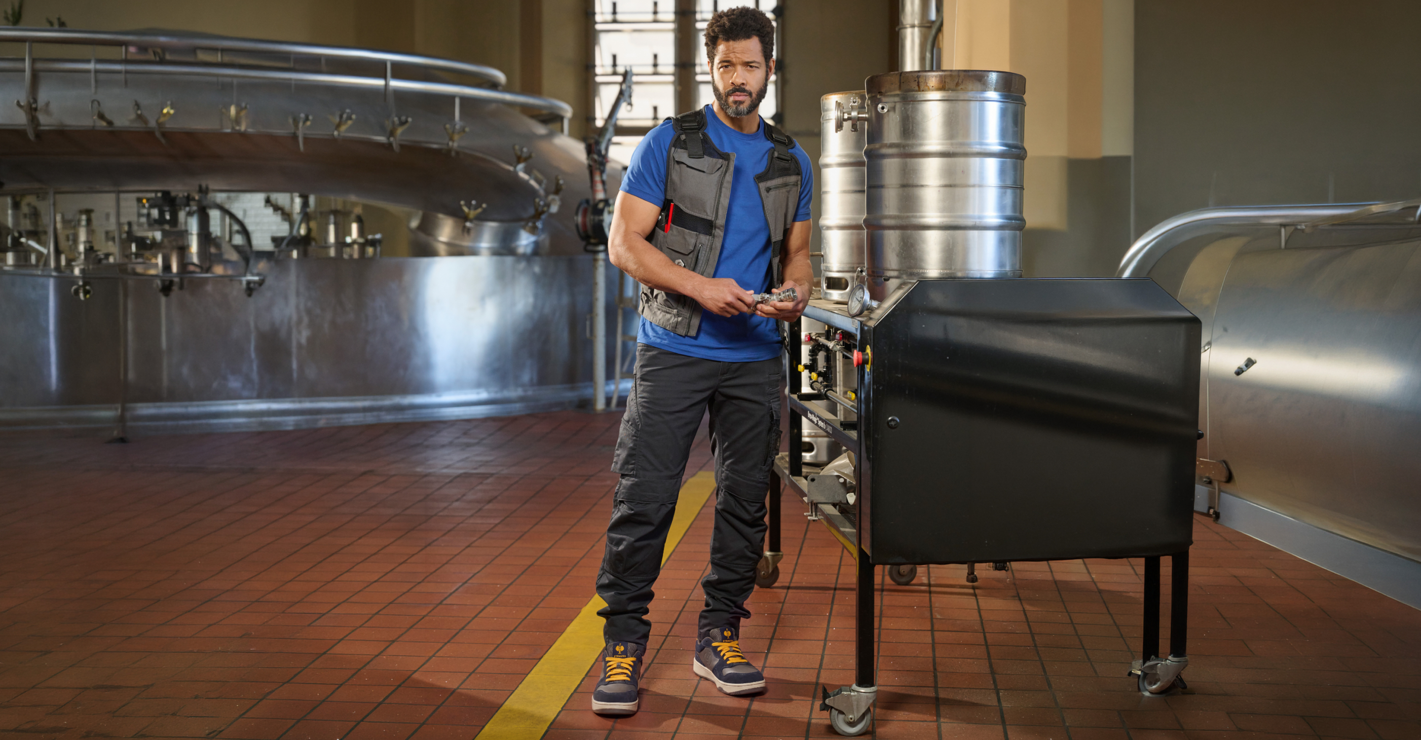 A man in work clothes stands in a brewery, holding tools beside a metal machine and a large keg, surrounded by stainless steel equipment and tiled floors.