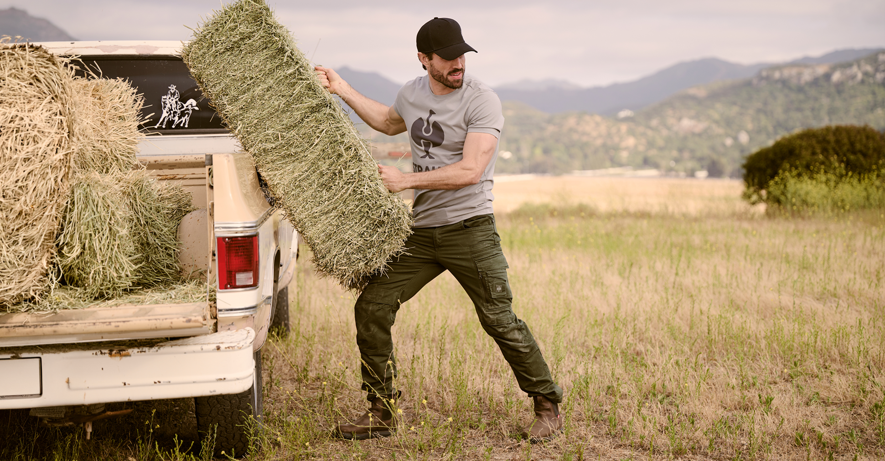 A man in a gray t-shirt and dark pants carries a large rectangular hay bale next to a white pickup truck loaded with hay, set in an open grassy field with mountains in the background.