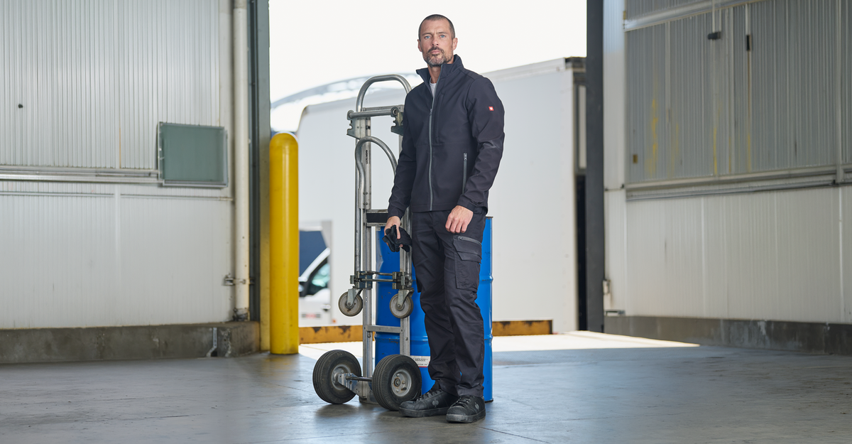 A man wearing a black work jacket and dark cargo pants stands next to a blue and silver industrial hand truck inside a warehouse.