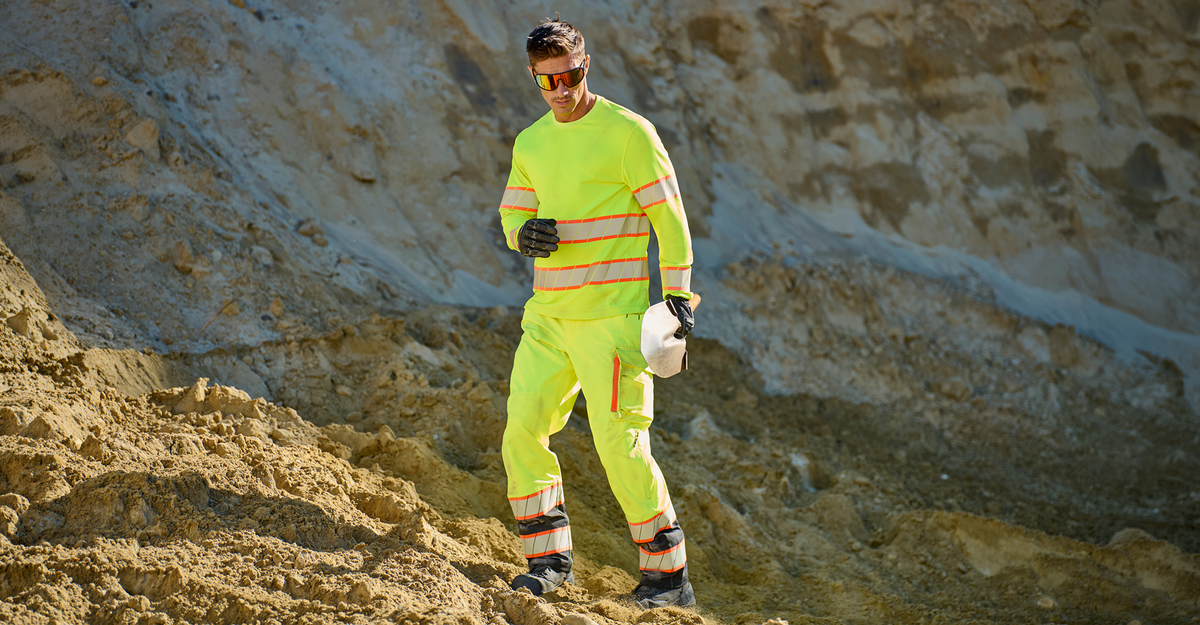 A man wearing a high-visibility neon yellow-green fire service uniform with reflective stripes, walking on a sandy, rocky outdoor terrain while holding a white hard hat.