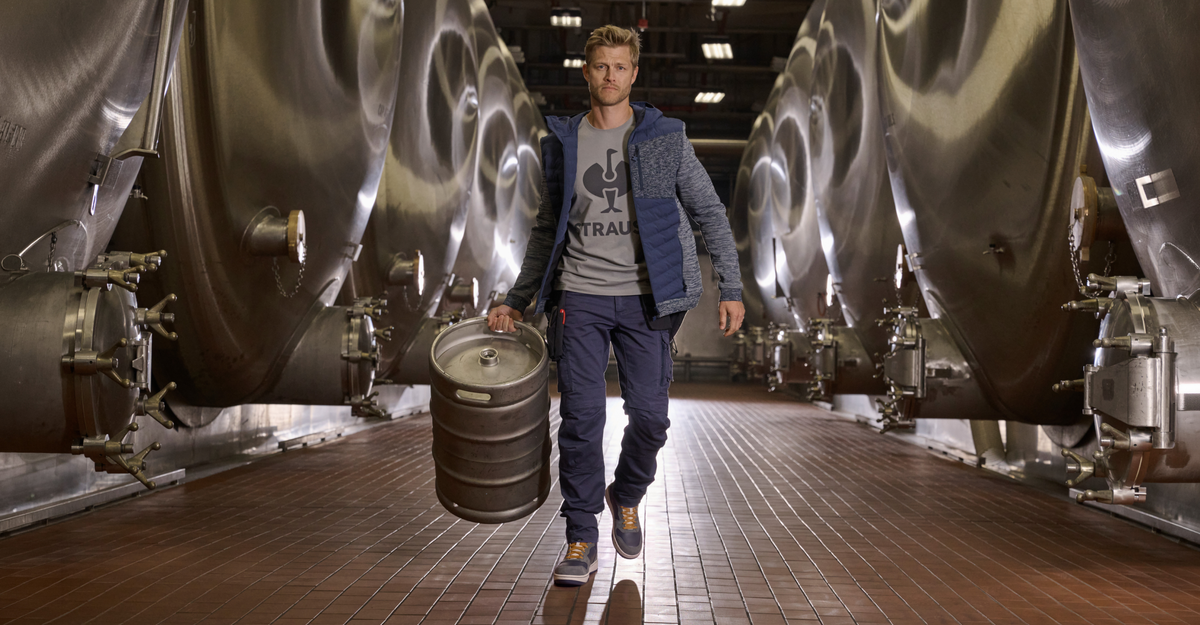 A man walks down a tiled aisle between large stainless steel fermentation tanks in a brewery, carrying a metal kegel. He is wearing a blue puffer jacket over a gray t-shirt with a logo, dark blue pants, and sneakers.