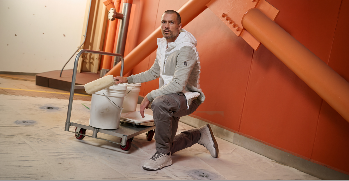 A man kneels on a floor covered with protective sheeting, holding a paint roller next to a white bucket and paint tray. He is wearing a light gray zip-up hoodie, gray pants, and white sneakers.