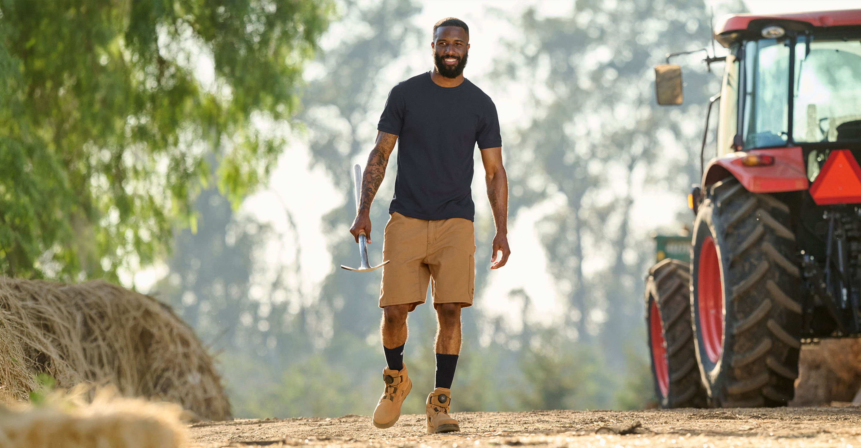 A man walking on a dirt path in a farm setting, wearing a dark gray t-shirt, tan shorts, and work boots, with a red tractor visible on the right.