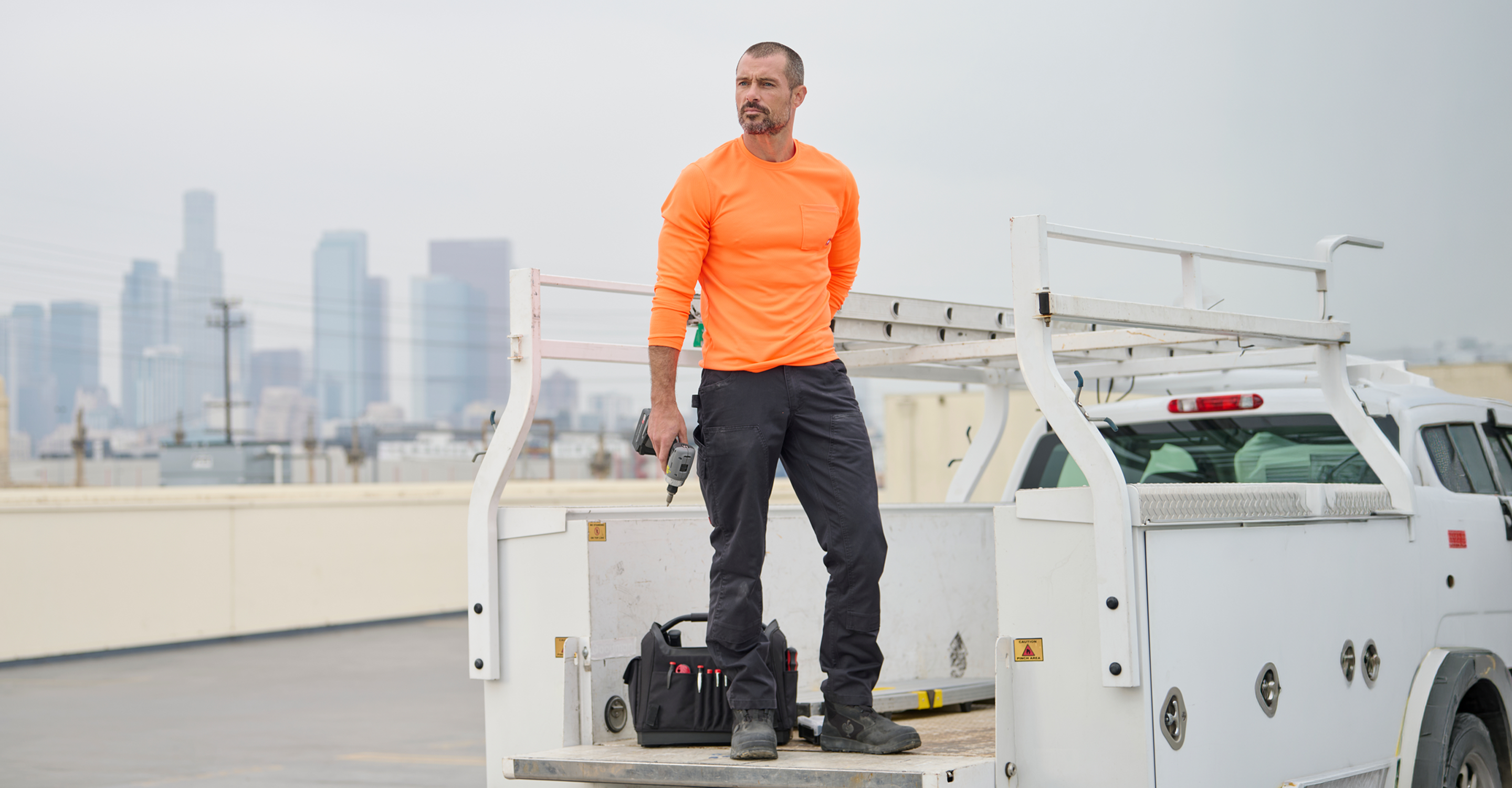 A man in an orange long-sleeve shirt and dark work pants stands in the back of a white utility truck on a rooftop, holding a drill. A black tool bag rests on the truck bed, and a city skyline is visible in the hazy background.