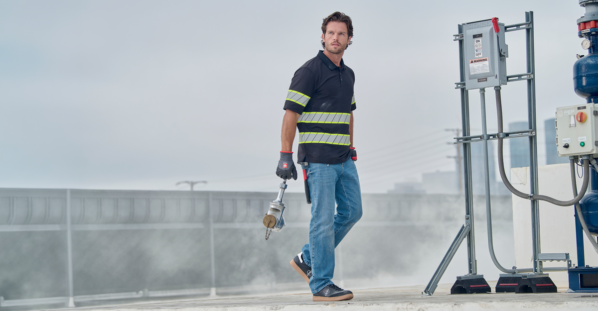 A man in a black and yellow reflective safety shirt, blue jeans, and work gloves walks on a rooftop while holding a cordless drill. Industrial equipment and metal piping are visible in the background under an overcast sky.