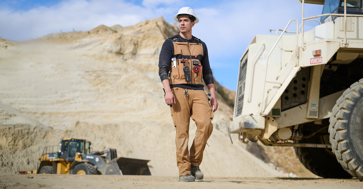 A construction worker in a hard hat and safety vest walks on a sandy site, with large construction vehicles and rocky terrain in the background.