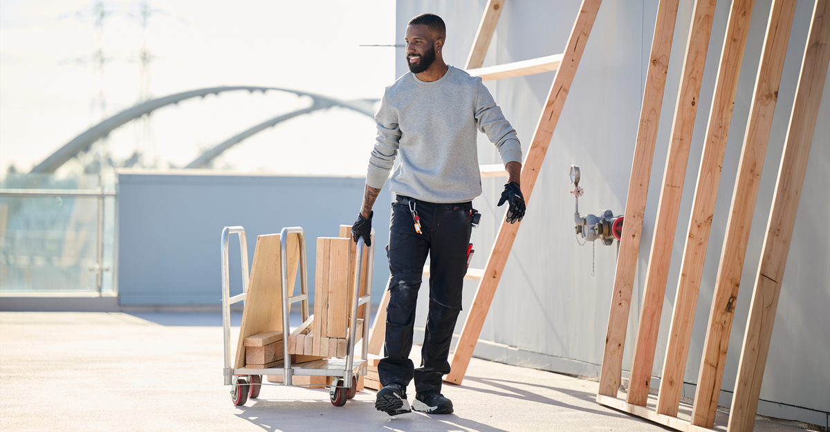 A man wearing a light gray sweatshirt, dark pants, and work gloves pushes a wooden pallet cart on a rooftop terrace next to exposed wooden framing.